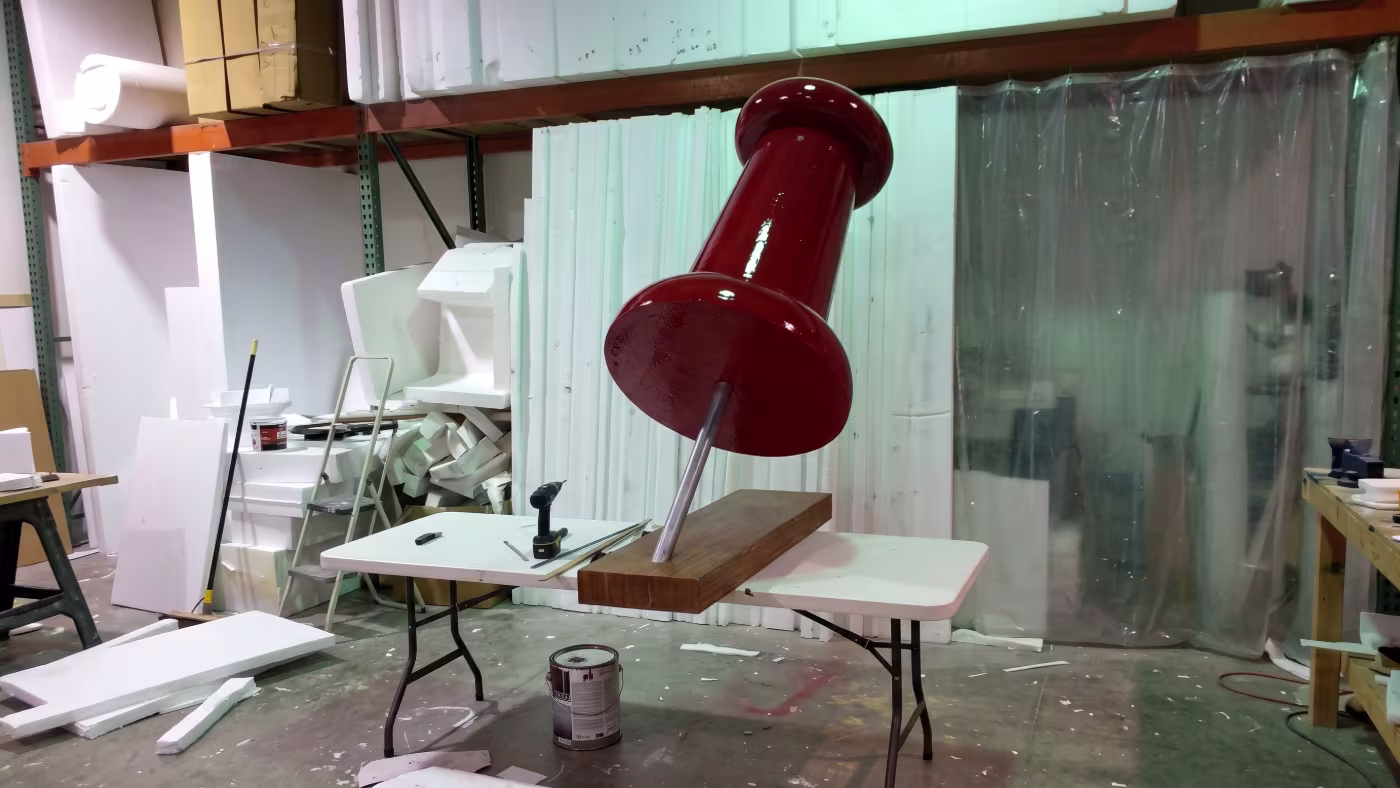 Chincheta gigante lacada en rojo, clavada en un tablero de madera, en la nave de fabricación